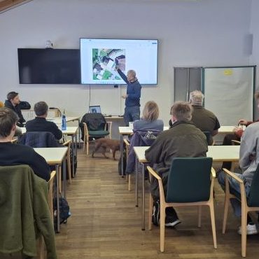 Vor einem modernen Lehrgebäude sitzen zwei junge Frauen und ein junger Mann mit Brille.